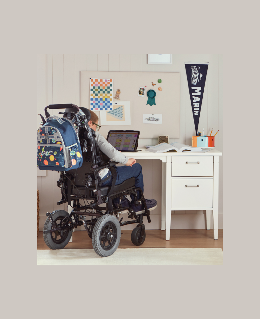 Image shows young, school-aged boy in a wheelchair sitting at a Pottery Barn Kids Morgan Desk - ADA in the corner of a kid's bedroom. His wheelchair has a Pottery Barn Kids Mackenzie Adaptive Backpack attached to it. The boy is reaching into the top desk drawer to grab a toy car.