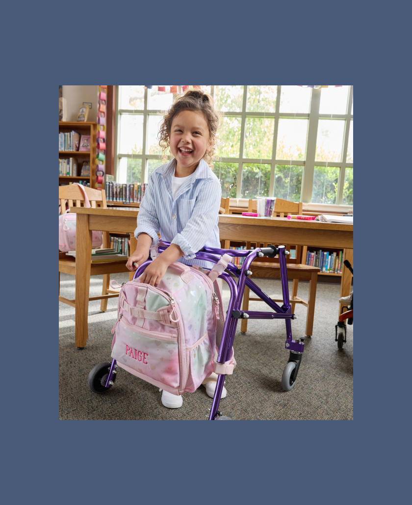 View from the side of young boy using a mobility device with Mackenzie Adaptive Backpack affixed to it.