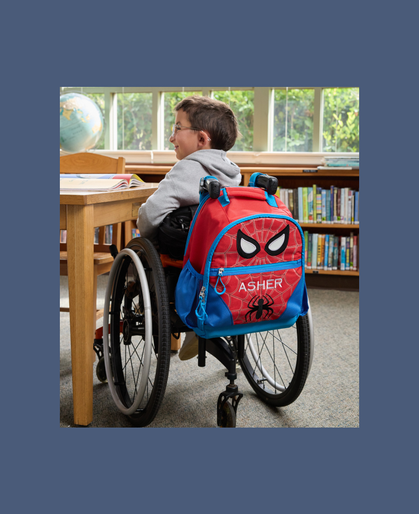 Image of young boy in wheelchair with Mackenzie Adaptive Backpack and Mackenzie Adaptive Lunch Box affixed to it. Next to him, a young girl using a mobility device with a Mackenzie Adaptive Backpack affixed to it.