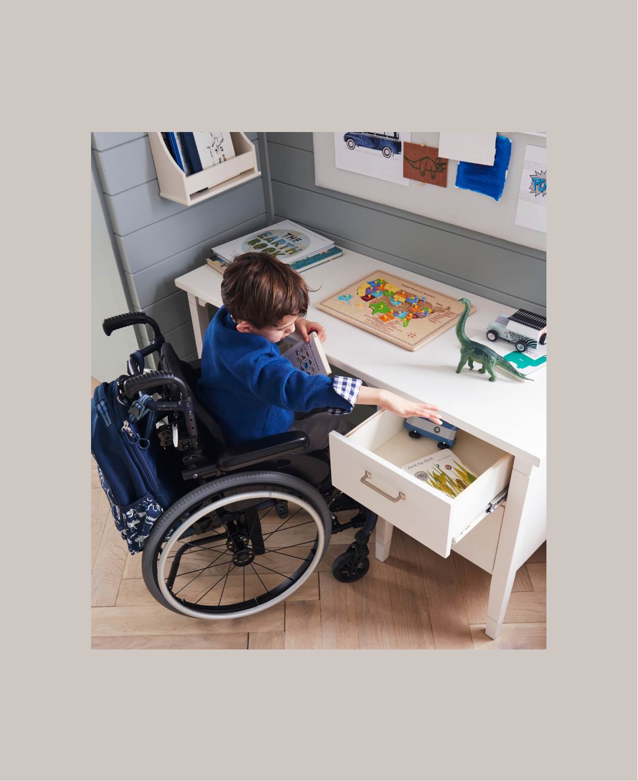 Image shows young, school-aged boy in a wheelchair sitting at a Pottery Barn Kids Morgan Desk - ADA in the corner of a kid's bedroom. His wheelchair has a Pottery Barn Kids Mackenzie Adaptive Backpack attached to it. The boy is reaching into the top desk drawer to grab a toy car.