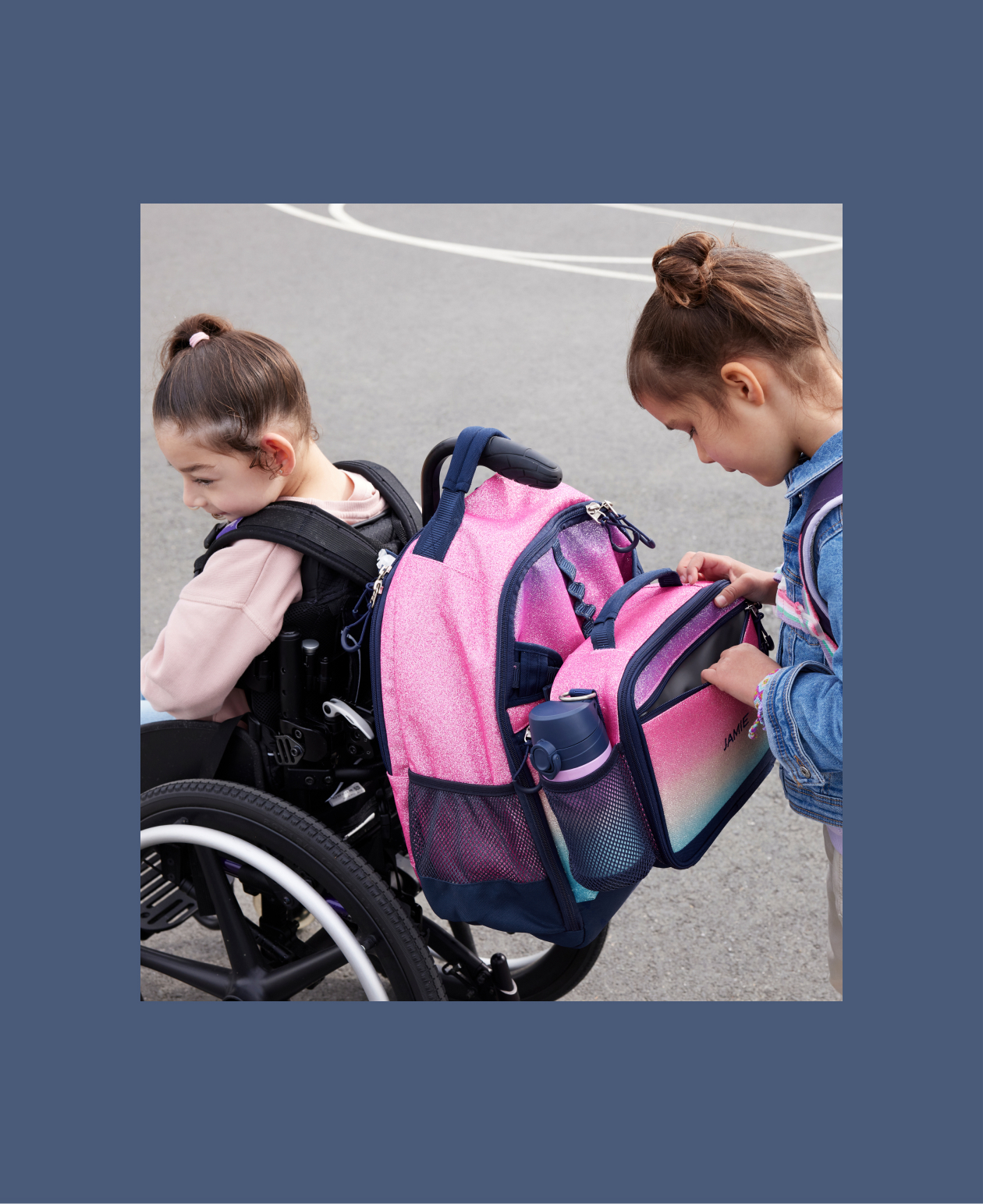 Image shows two young girls on a school blacktop. One girl sits in a wheelchair with a Pottery Barn Kids Mackenzie Adaptive Backpack in Ombre Glitter print attached to it. Attached to the backpack is a Pottery Barn Kids Mackenzie Adaptive Lunch Box in Ombre Glitter print. The second young girl looks into the backpack to retrieve something.