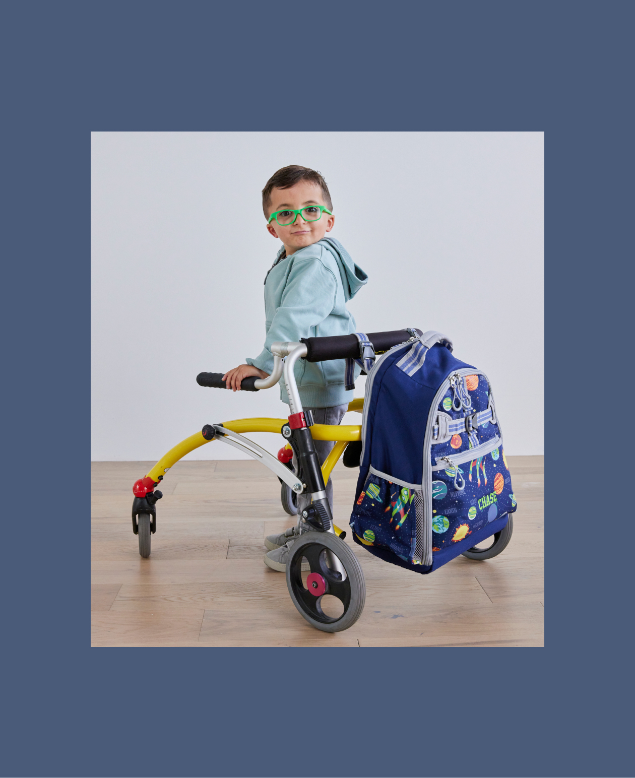 Image shows a different young boy standing with a walker. Attached to the walker is a Pottery Barn Kids Mackenzie Adaptive Backpack in Solar System print and embroidered with a name.
