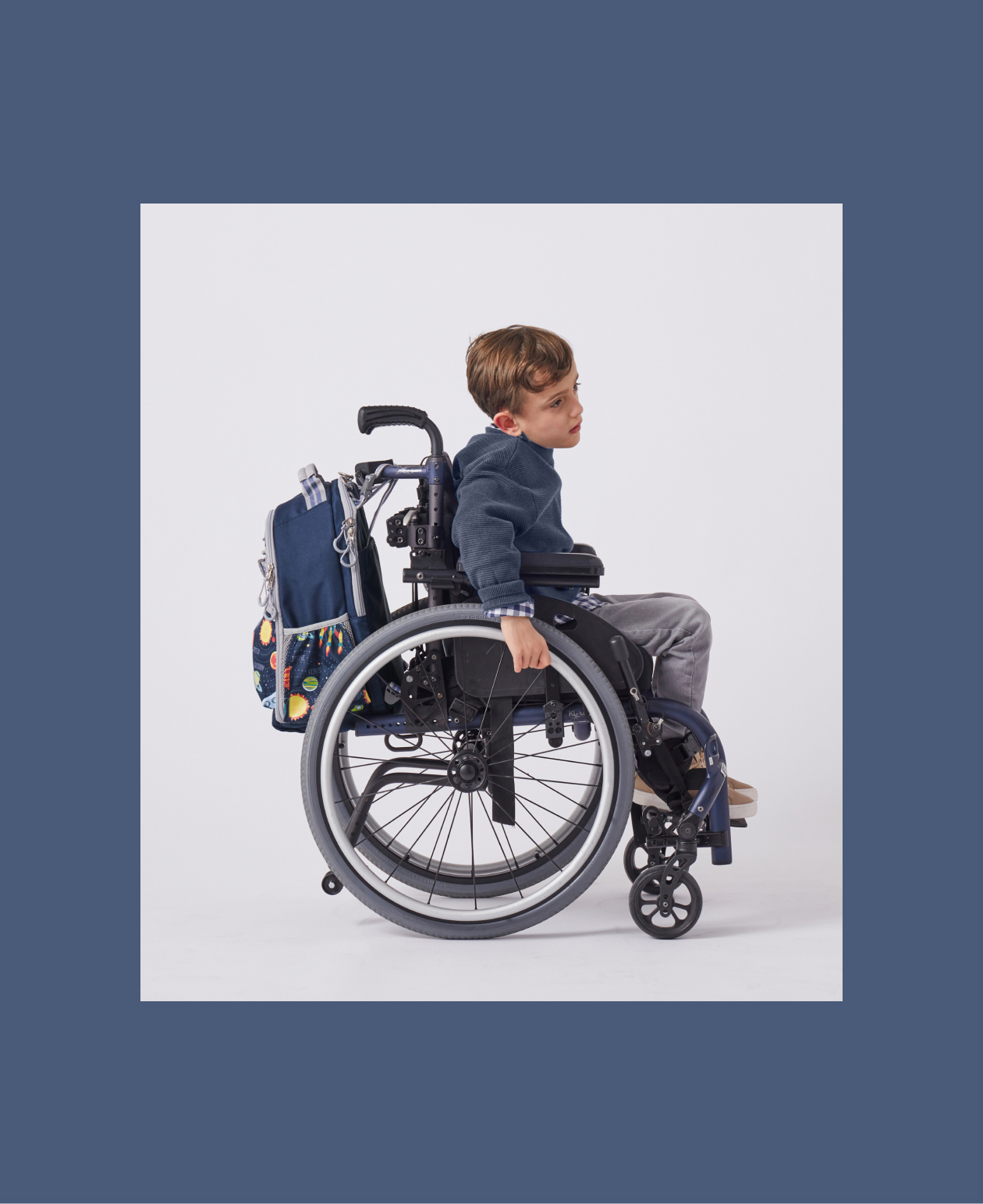 Image shows the side profile a young boy sitting in a wheelchair. Attached to the wheelchair is a Pottery Barn Kids Mackenzie Adaptive Backpack in Solar System print and embroidered with a name.