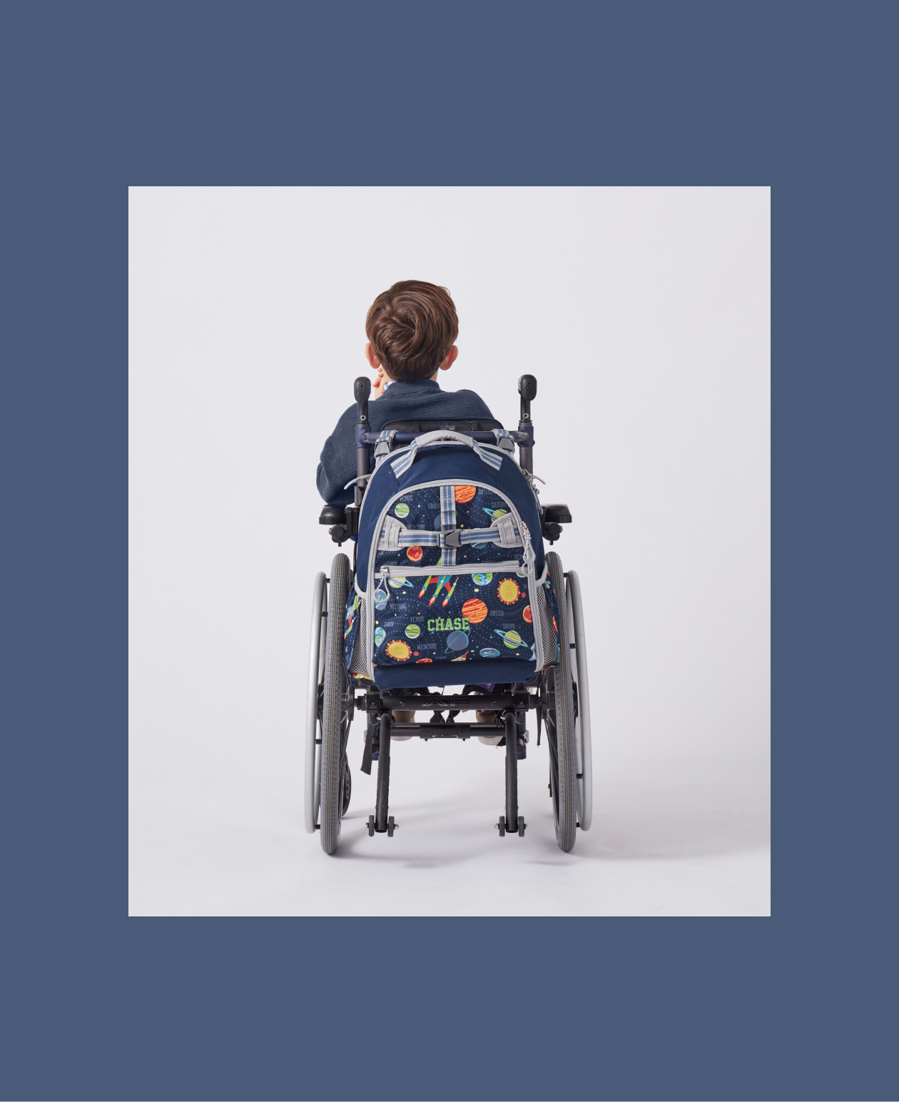 Image shows the back profile a young boy sitting in a wheelchair. Attached to the wheelchair is a Pottery Barn Kids Mackenzie Adaptive Backpack in Solar System print and embroidered with a name.