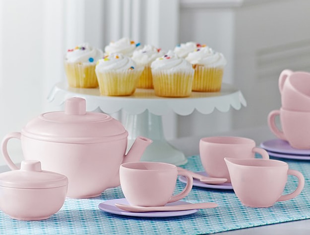 Matching pink tea set in front of a white platter of frosted cupcakes