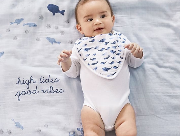 Baby laying down wearing a white onesie and drool bib with blue whales on it