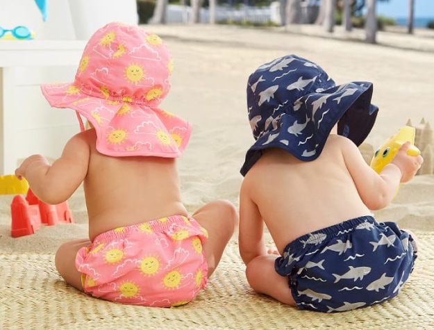 two young children playing with toys on beach mat, wearing pink sun-themed and blue shark-themed diaper covers and hats