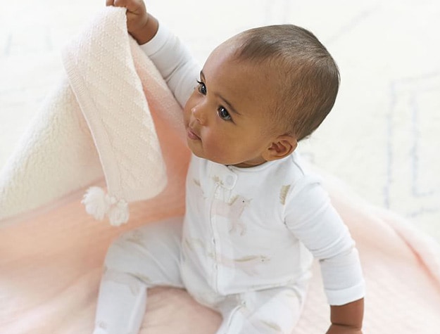 Baby in unicorn onesie sitting on a pink sweater knit baby blanket.