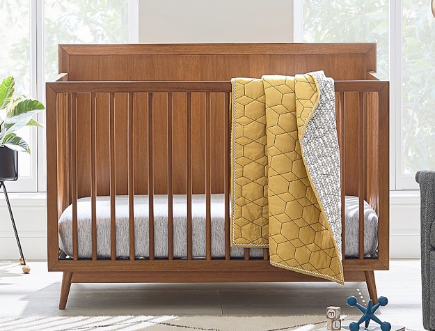 West Elm x pbk Mid-Century 4 in 1 Convertible Crib in acorn with yellow blanket draped over side.