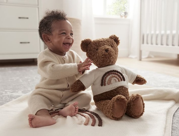 A baby on a blanket playing with an NAACP Give Back Teddy Bear Plush in a nursery.