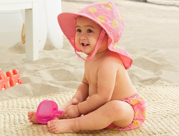 A baby wearing matching sunshine bottom and hat holding a beach toy sitting on a blanket at the beach.