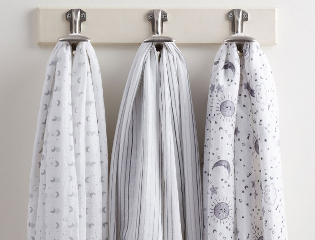 Three organic cotton muslin blankets, part of the Skye Organic Swaddle Set, hanging on individual hooks on a wall.