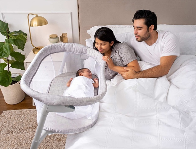 A couple looks into a portable bassinet that holds their baby at their bedside.