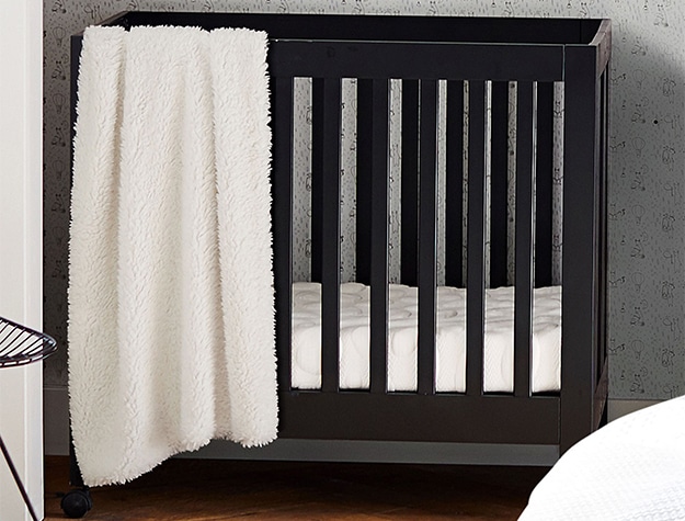 A black mini crib in the corner of a room with adjustable height and a white blanket draped over the side. 