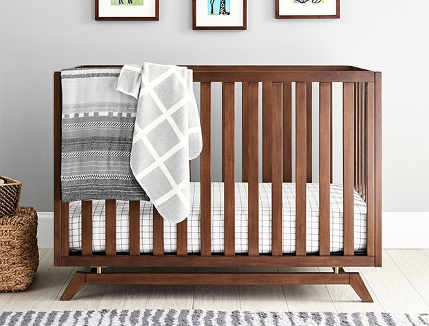 A wooden convertible crib sitting against a wall in a nursery with a gray blanket draped over the side.
