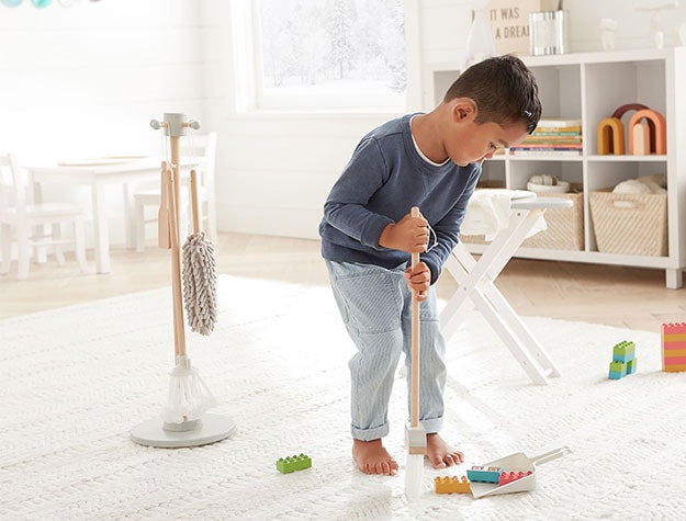 A child plays with wooden cleaning tools on a white rug.