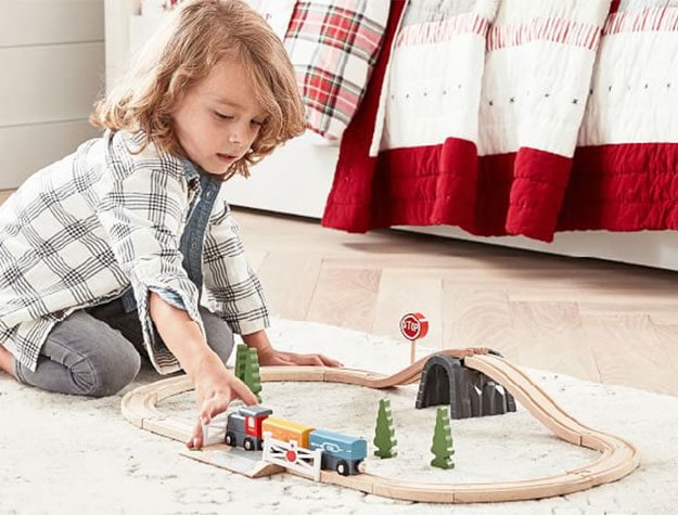A child plays with a wooden train set on a neutral rug.