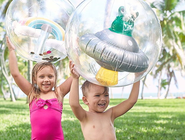 Two children playing with whimsical beach balls outside in sunshine and green grass.