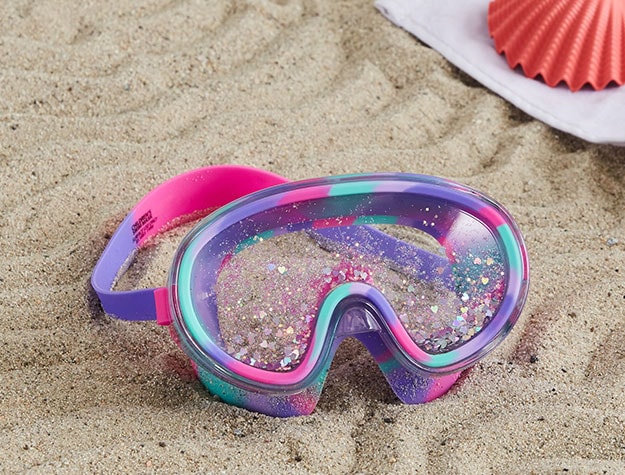 Pink and purple glittery swim goggles sitting in the sand.