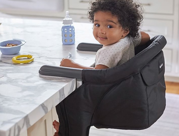 Baby holds a spoon in a portable table chair attached to a counter holding a spoon.