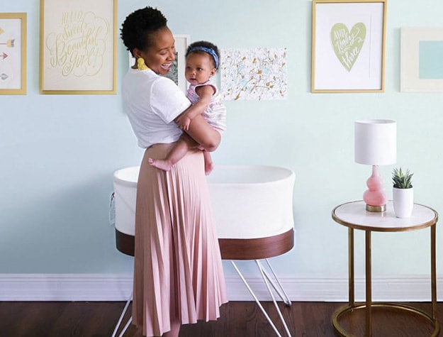A mother holds a baby in front of a bassinet in a stylishly decorated room.