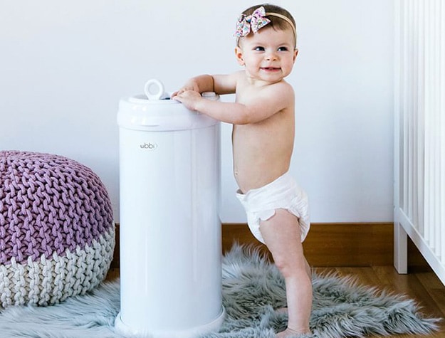 Baby wearing a bow standing next to a diaper pail in a nursery.