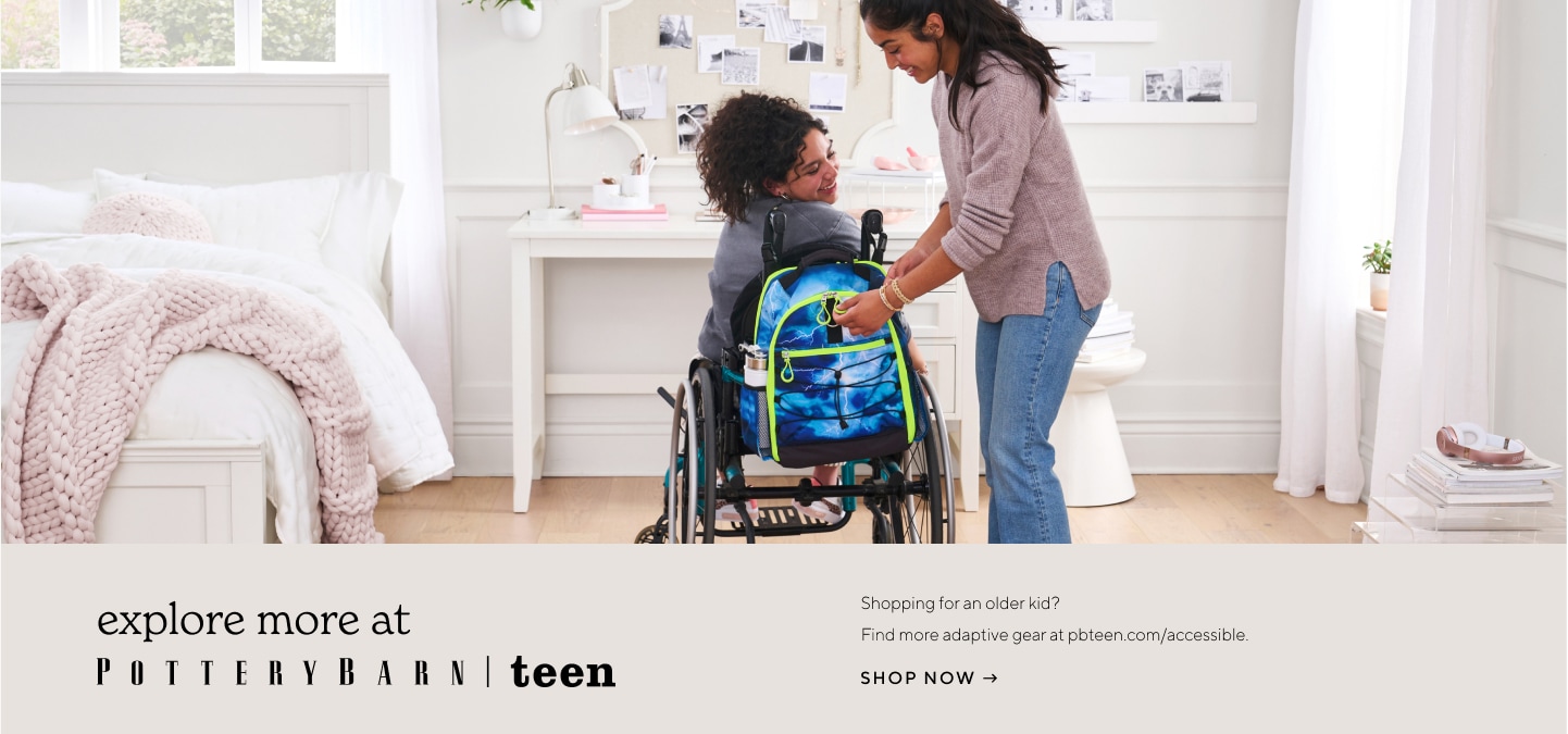 Image shows two teen girls in a white and pink teen girl bedroom. One girl sits in a wheelchair with a Pottery Barn Teen Gear-Up Adaptive Backpack in Storm print attached to it. The other girl stands next to her and helps to unzip the backpack.