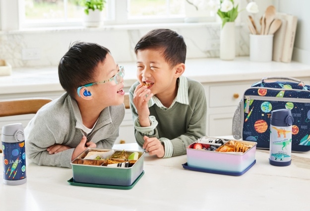 Two young boys sit at the kitchen counter eating lunch, showcasing their Pottery Barn Kids lunch boxes, water bottles, and all-in-one bento boxes.