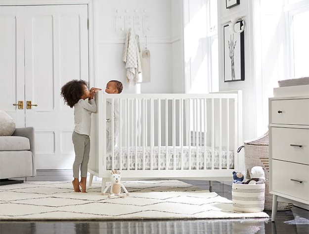 A young girl peeks over the edge of a crib to see a toddler standing inside the crib. A young girl peeks over the edge of a crib to see a toddler standing inside the crib.