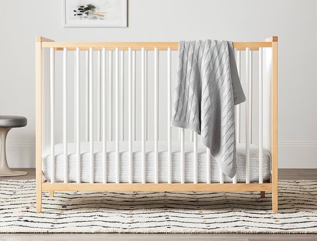 A light brown crib with white slats and a gray blanket hanging on its side. A light brown crib with white slats and a gray blanket hanging on its side.