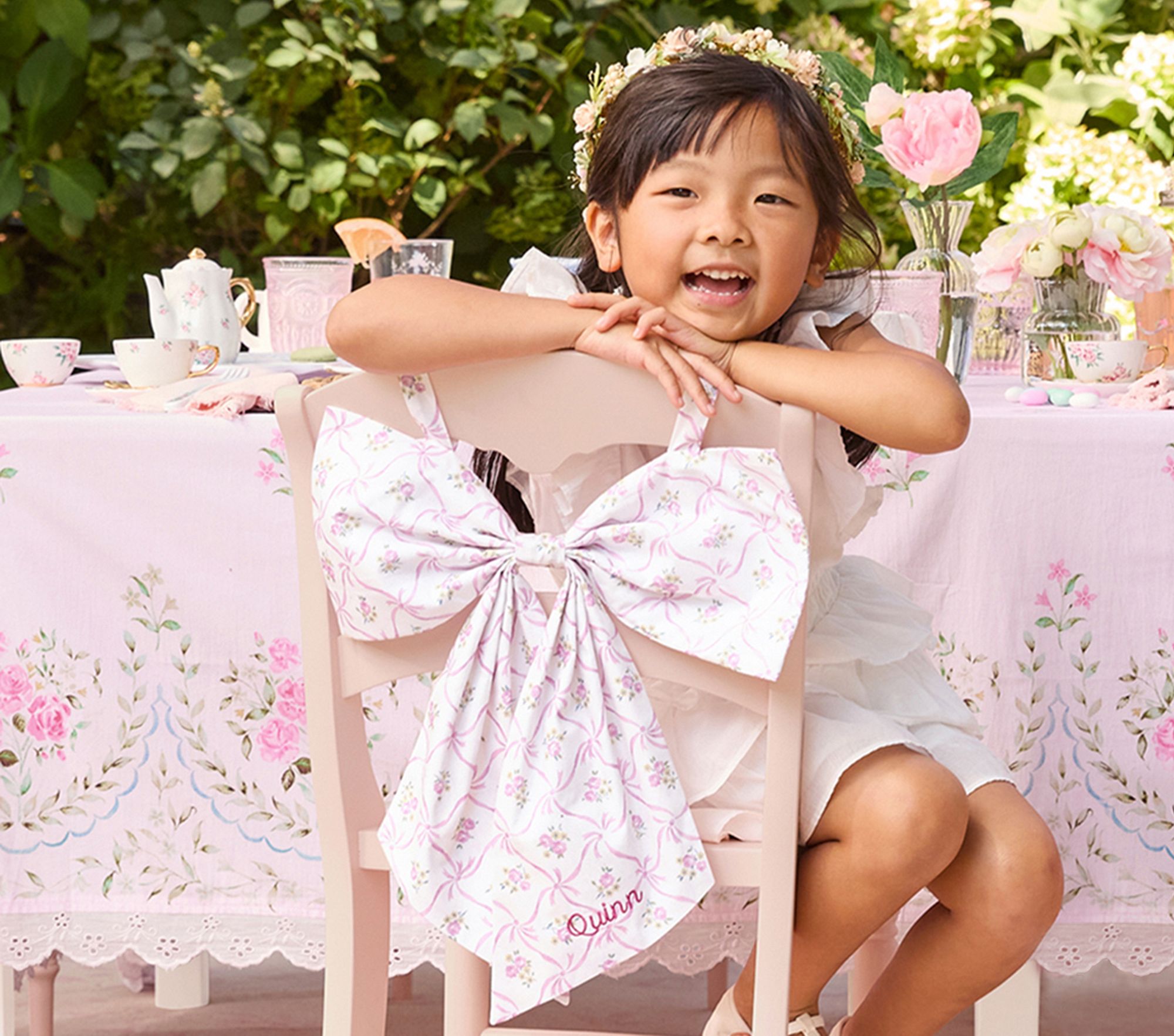 Child wearing a dress with pink floral bow chairbacker tied to chair.
