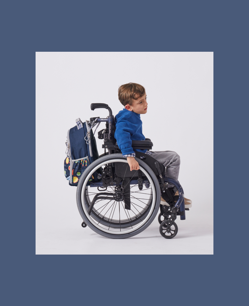 View from the side of young boy in wheelchair with Mackenzie Adaptive Backpack affixed to it.