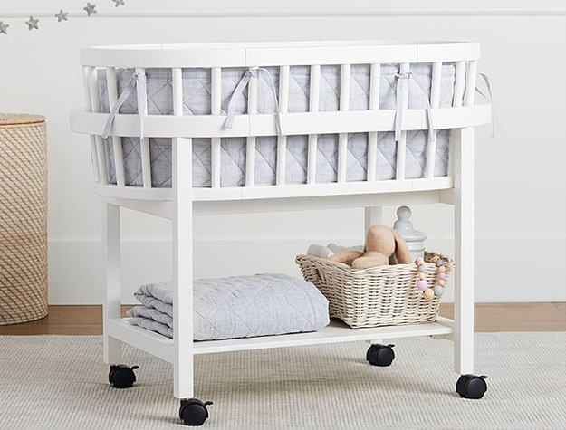 A white bassinet with a basket of toys and a folded blanket beneath it.