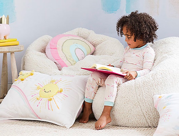 Toddler reading a book in a bean bag chair next to bright throw pillows
