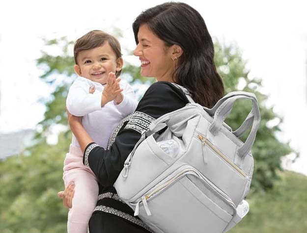 A woman holding a baby and wearing the Skip Hop mainframe wide open backpack diaper bag