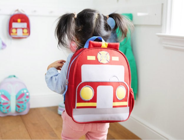 A child with pigtails wears a firetruck backpack monogrammed with the name Kate.