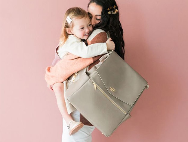 Mother holding baby and wearing the Freshly Picked Classic Diaper Bag.