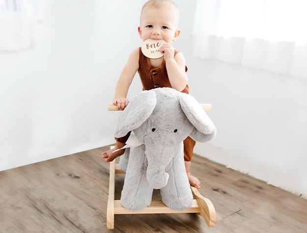 Baby sitting on gray elephant critter nursery rocker holding plaque reading one year.