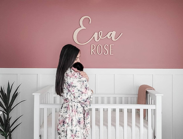 Mother holding baby in front of crib with wall letters reading Eva Rose in background. 