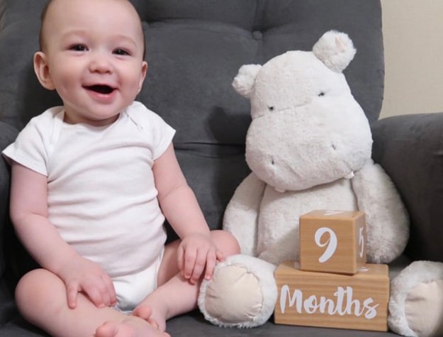 Smiling baby sitting on a plush chair next to a gray hippo plush and blocks reading 9 months.