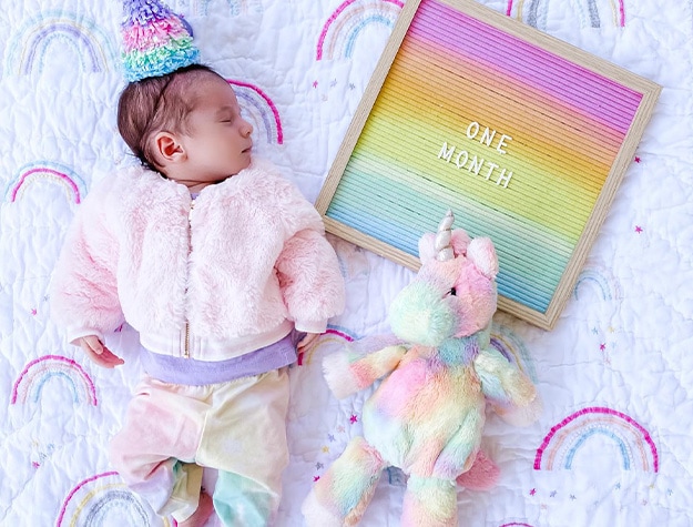 Sleeping baby with stuffed unicorn, rainbow bedding and felt letterboard reading one month.