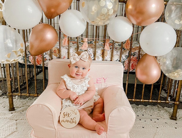 Baby in an Anywhere Chair surrounded by balloons holding a placard reading six months.