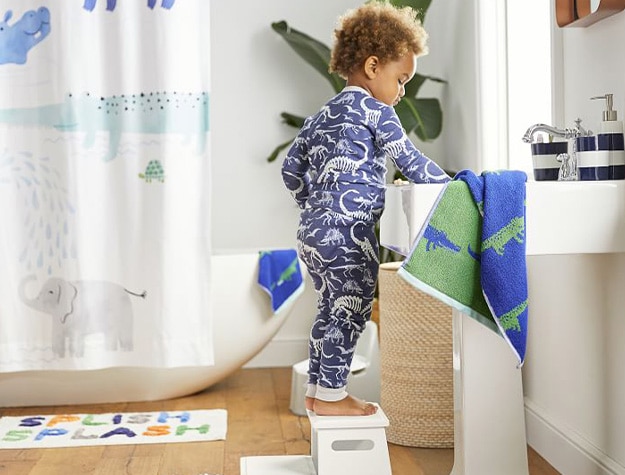 Child in dinosaur themed pajamas standing on stepping stool to reach a bathroom sink.