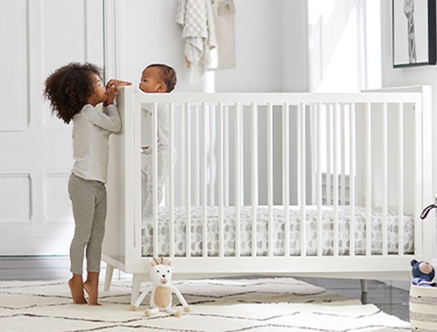Child looking over crib to play with baby sibling in a white themed nursery.