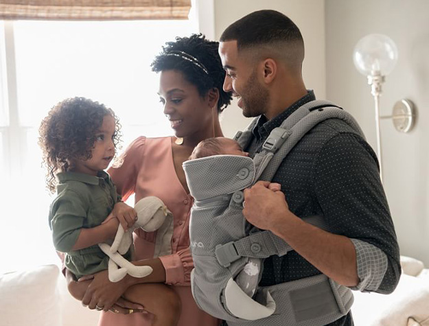 Mother holding young child with stuffed bunny and father holding baby strapped into Nuna CUDL baby carrier.