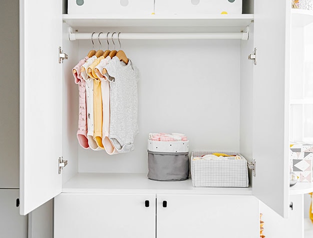 Baby clothes hanging in white closet with storage bins.