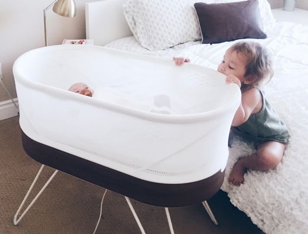 Happiest Baby SNOO Smart Sleeper Bassinet on side of bed with baby inside and toddler looking down at baby.