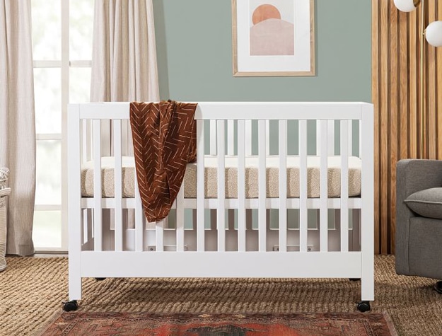 White Babyletto Maki Folding Crib with wheels in bedroom.