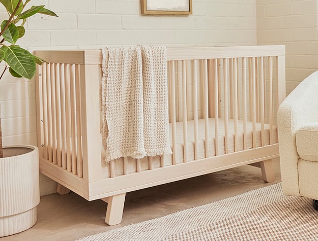 Washed natural wood Babyletto Hudson 3 in 1 Convertible Crib with tan blanket draped over side next to plant and chair.