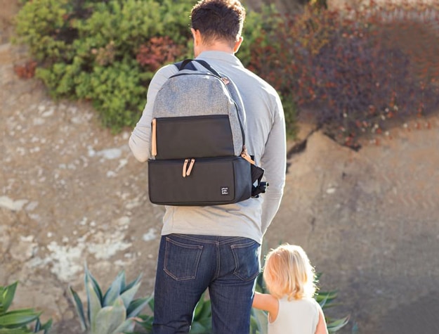 A parent walking their child outside wearing a Petunia Pickle Bottom Axis Backpack containing daycare essentials.